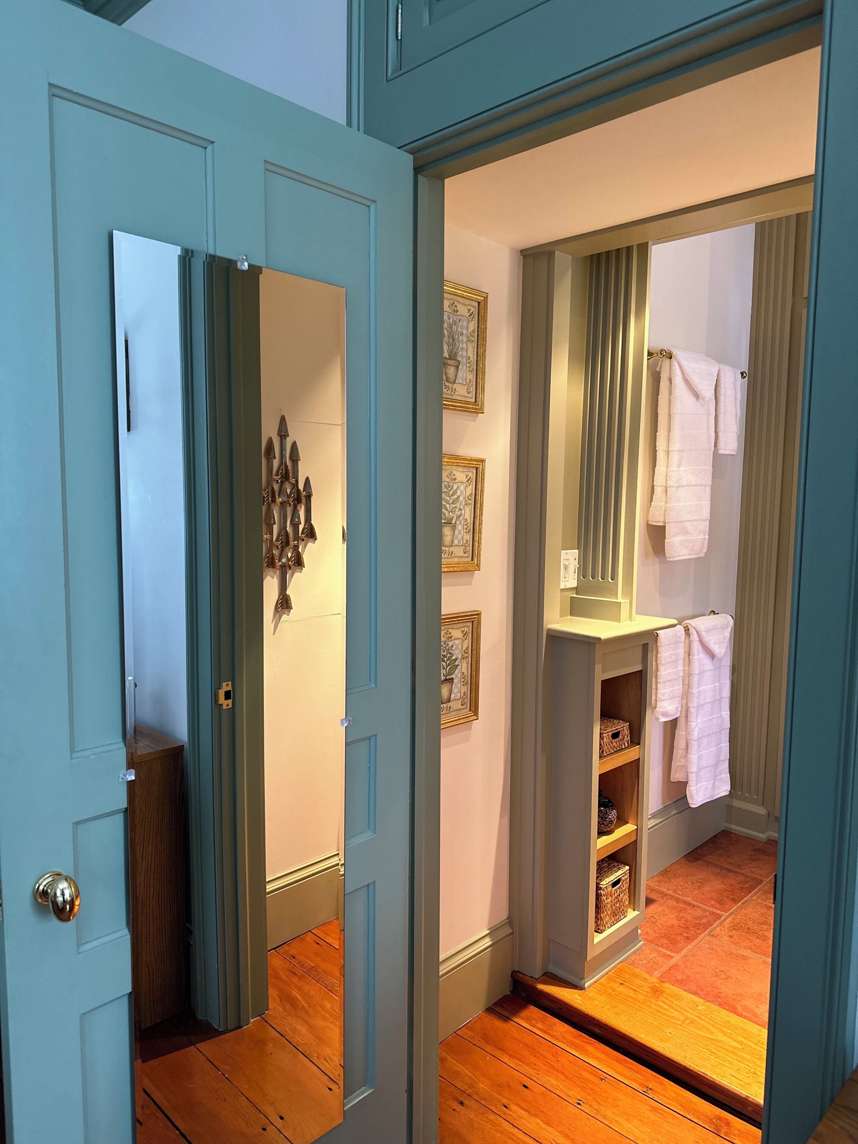 Un couloir doté d’un plancher en bois, d’une porte bleue et d’une salle de bain équipée d’un porte-serviettes et d’étagères.