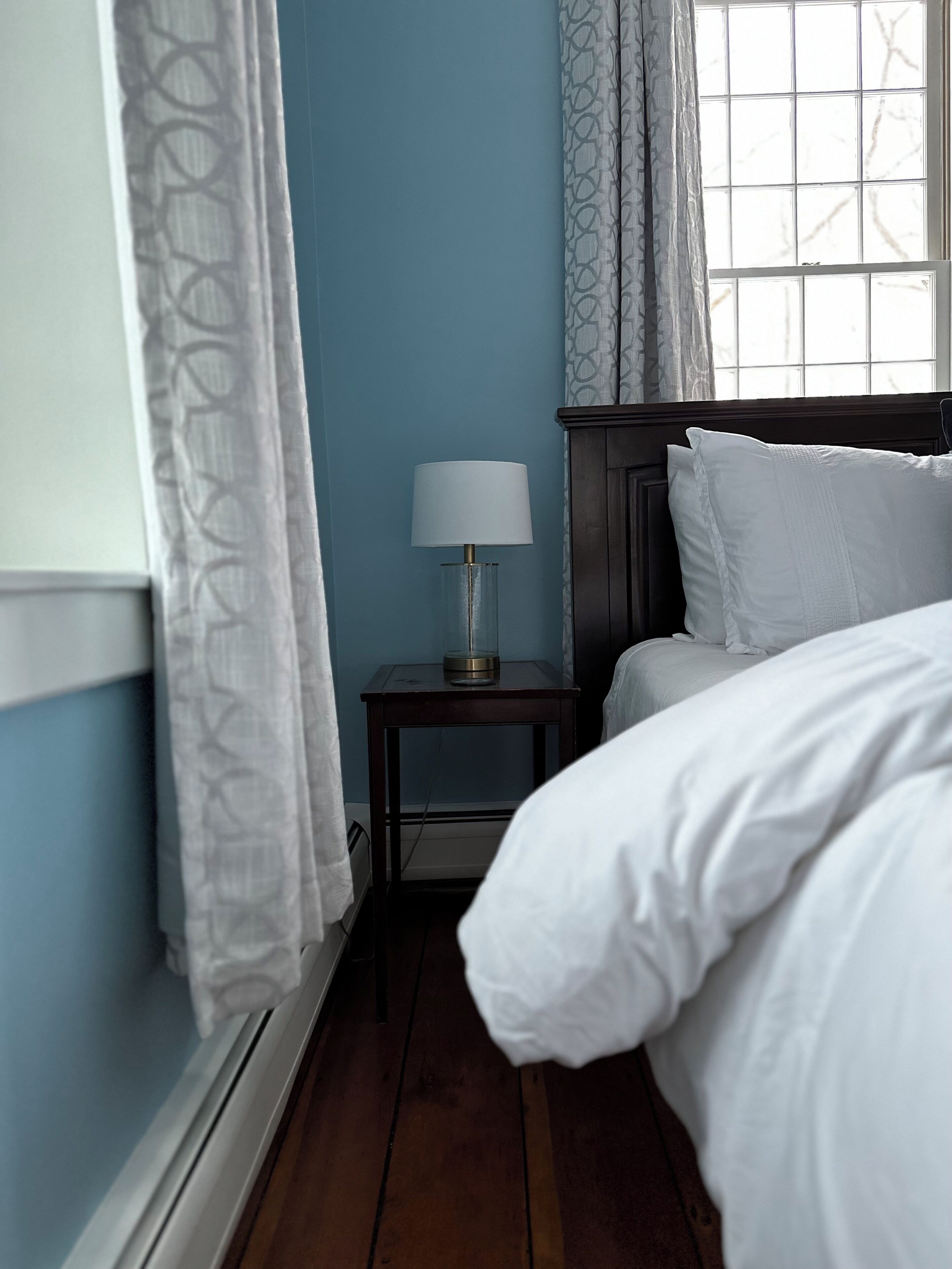 A bedroom with a wooden bed, a bedside table with a lamp, a window with curtains, and a blue wall.
