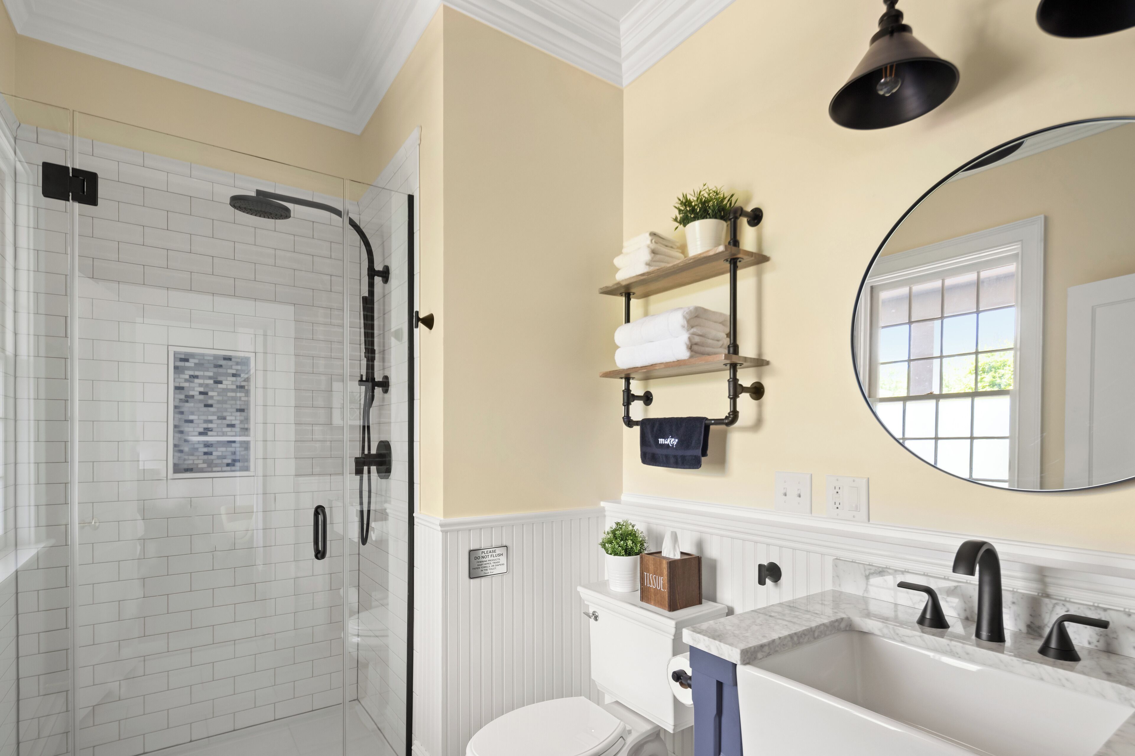A bathroom with a glass-enclosed shower, a white toilet, a sink with a marble countertop, and a shelf with towels and a plant.