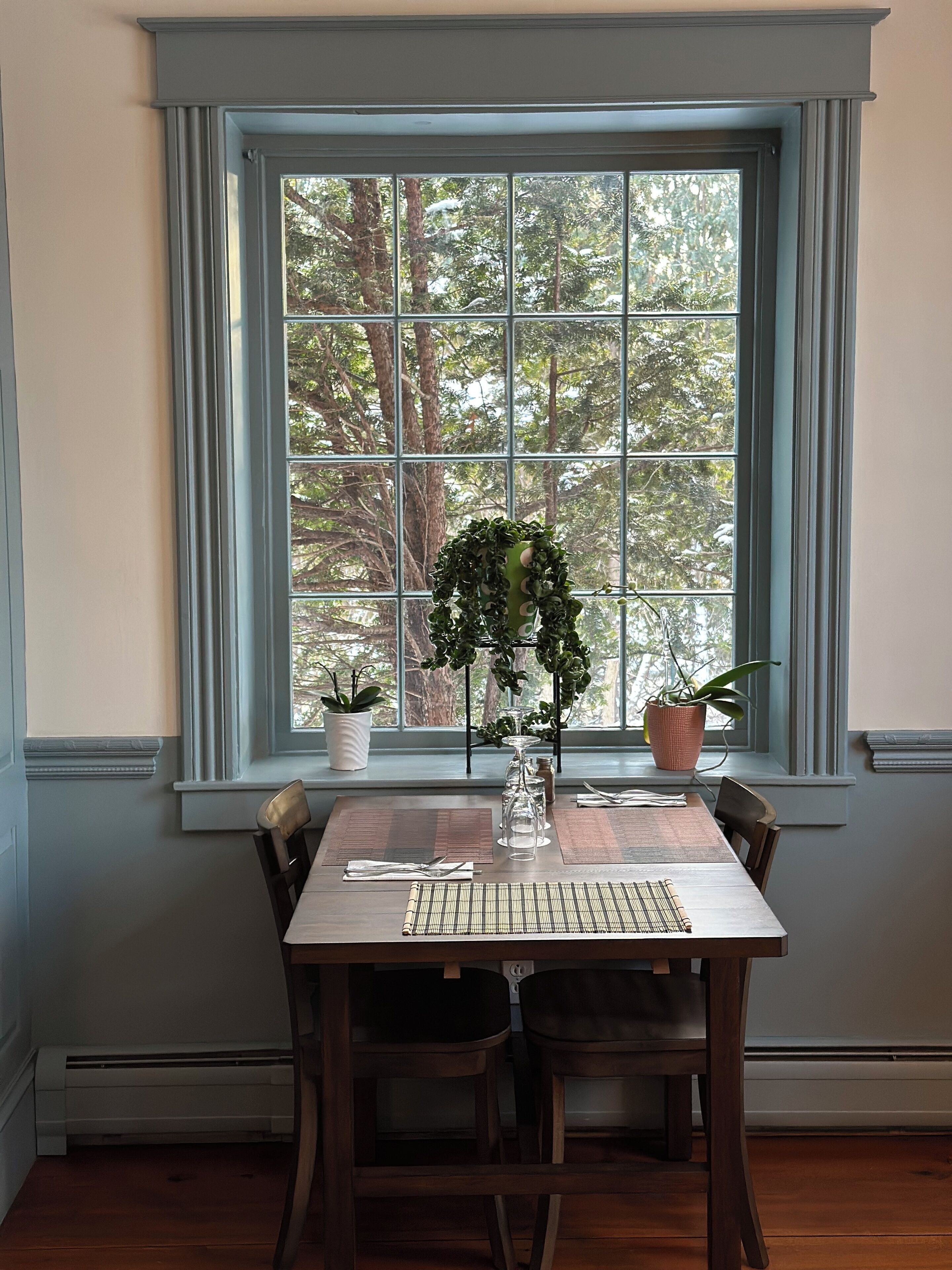 A dining table set for four with chairs, a window with a view of trees, and two potted plants on the windowsill.