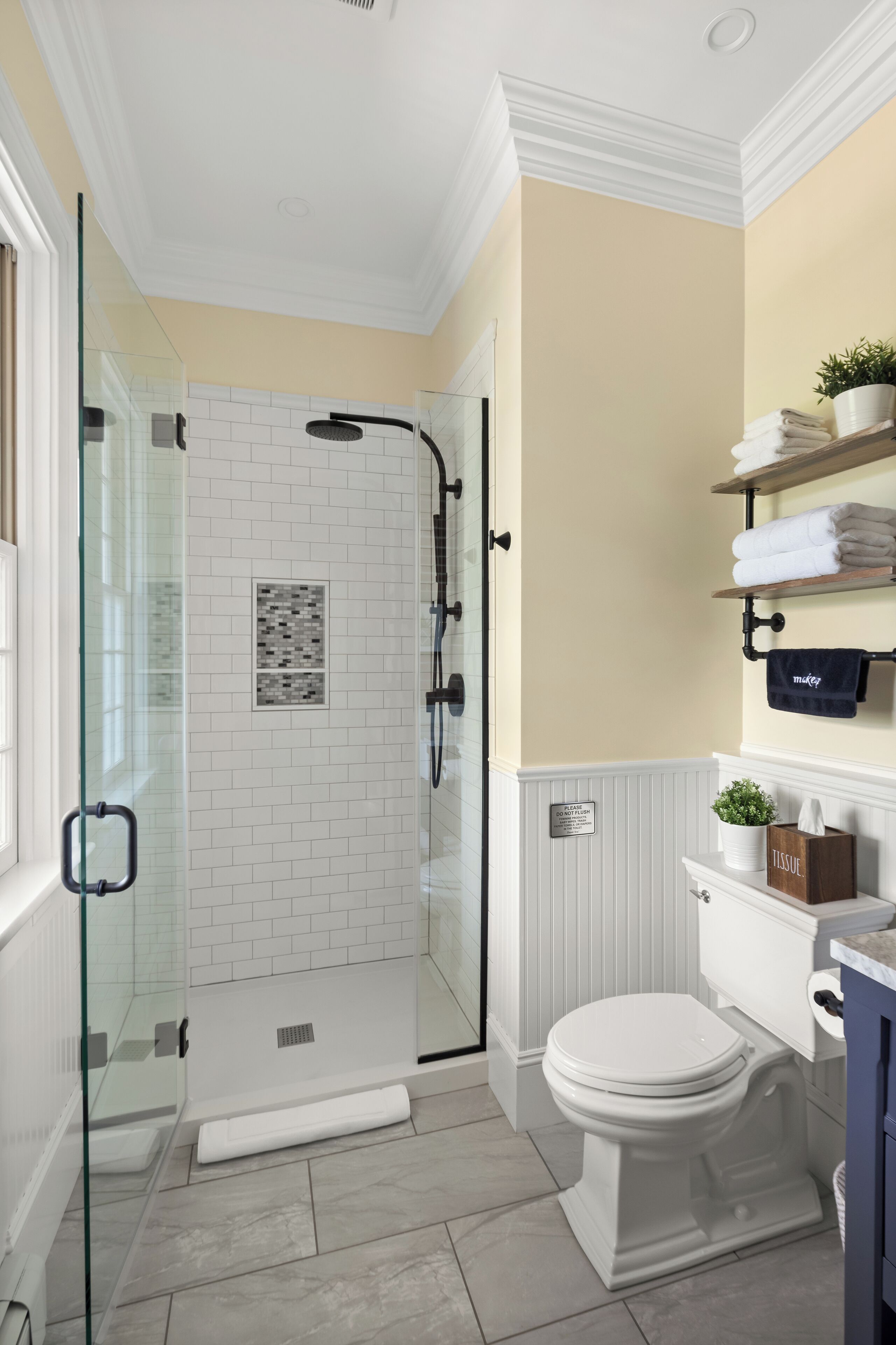 Un baño con ducha de vidrio, paredes cubiertas de azulejos blancos, un inodoro blanco y una pared color beige con un perchero y una planta en maceta.
