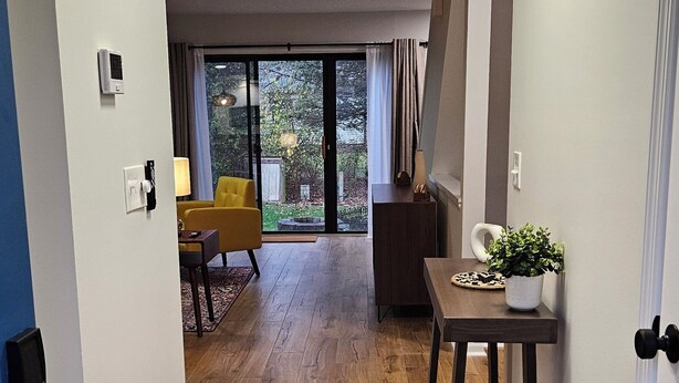 A hallway with wooden flooring leading to a living area with a yellow chair, a table with a plant, and a view of the outdoors through large windows. - N. Columbus Townhome with 3bd, 2.5ba, Centrally Located! (Columbus)
