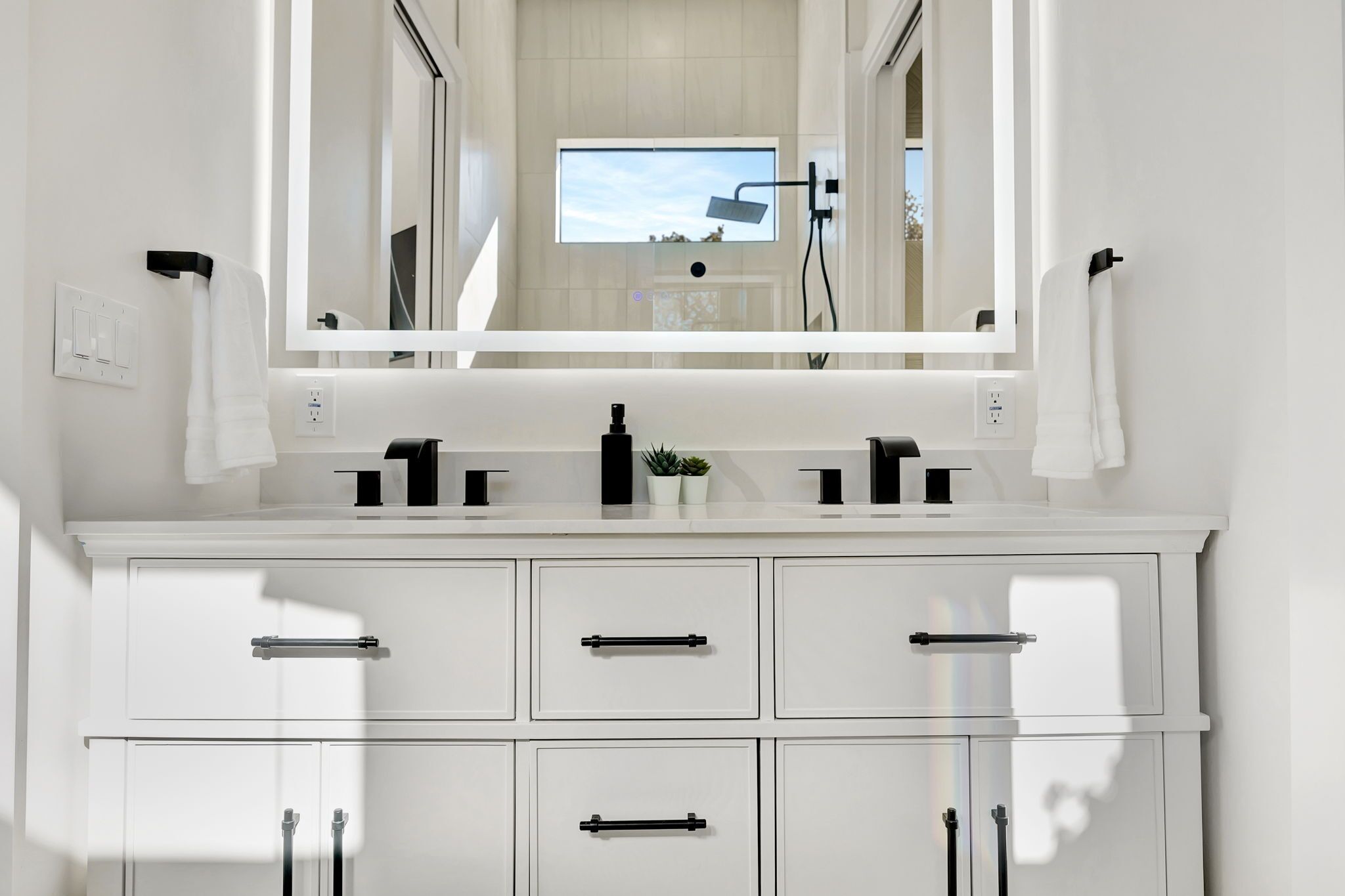 Un baño moderno con un espejo grande, dos lavamanos, una planta en maceta y una ventana.