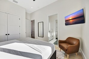 A modern bedroom with a large bed, a brown leather chair, a wall-mounted TV, and a door leading to another room. - New Upscale Retreat Downtown Dunedin w/ Balcony! Sleeps 4 (Dunedin)