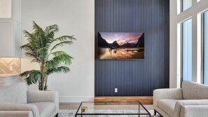 A modern living room with a large flat-screen TV mounted on a dark wall, a potted plant, and two sofas facing a glass coffee table.