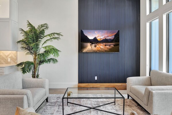 Una sala de estar moderna con un televisor de pantalla plana grande montado en una pared oscura, una planta en maceta y dos sofás orientados hacia una mesa de centro de vidrio.