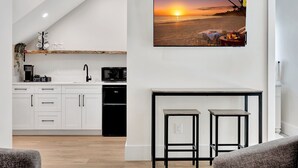 A modern kitchen with white cabinets, a coffee bar, and a microwave. There is a dining area with two bar stools and a table. A large picture of a beach sunset is displayed on the wall.
