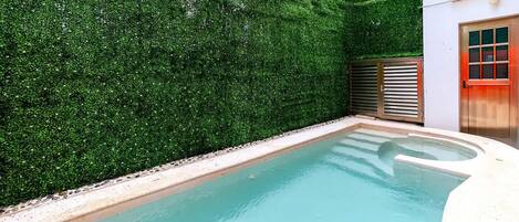 Une piscine entourée d’une haie, une porte avec un cadre rouge et un petit bâtiment doté d’une fenêtre.
