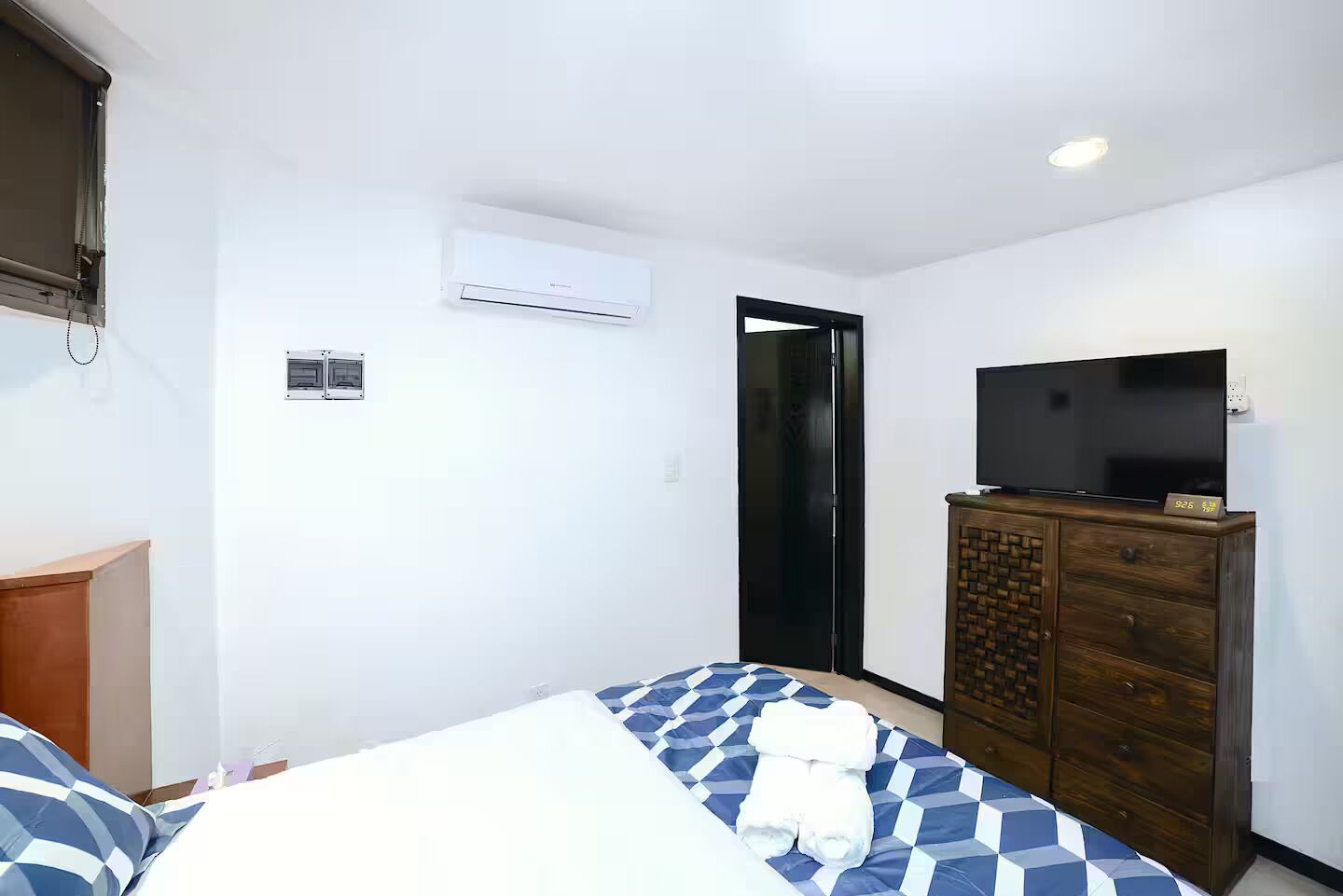 Una camera d'albergo con un letto, una cassettiera in legno, una televisione e un condizionatore d'aria.