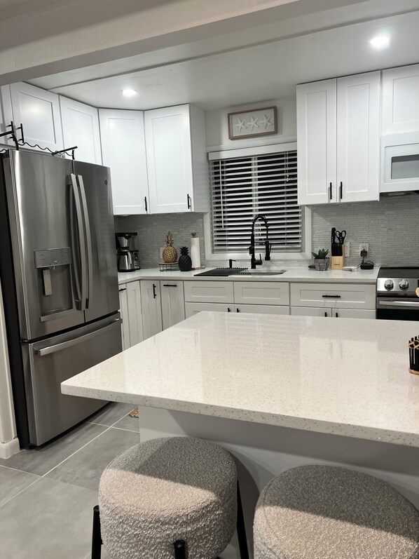 A modern kitchen with white cabinetry, a stainless steel refrigerator, a large island with stools, and a window with blinds.