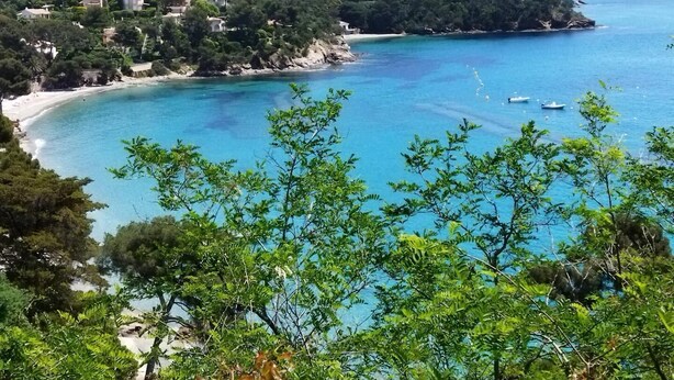 Une vue côtière comprenant une plage, une eau d’un bleu limpide et une colline boisée parsemée de bâtiments.