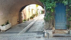 Une rue pavée, bordée d’une arche en pierre, avec une porte bleue et de la verdure.