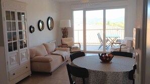Un salon avec un canapé beige, une table à manger avec des chaises, un meuble et une grande fenêtre donnant sur l’extérieur.