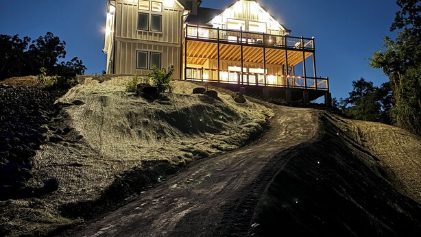 Une maison à deux étages dotée d’un balcon, située sur un chemin de terre avec une pente abrupte.