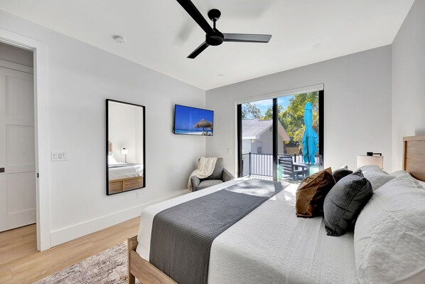 Un dormitorio moderno con una cama grande, un ventilador de techo, un televisor y un balcón con vistas a árboles y un edificio.
