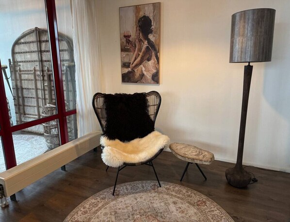Un salon moderne avec un fauteuil à armature métallique noire, un tapis rond, un petit pouf, une lampe sur pied et une grande fenêtre donnant sur un intérieur au style rustique.