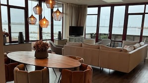 A modern living room with a round dining table, beige sofas, and a large window overlooking a body of water.