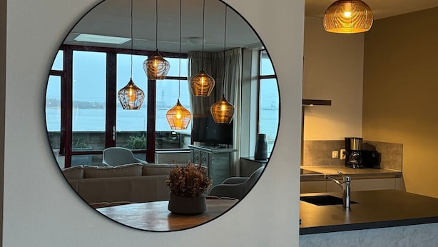 A modern kitchen with a round mirror reflecting a living room with a sofa, a coffee machine, and pendant lights.
