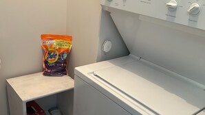 Een kleine wasruimte met een voorlader wasmachine, een plank met een zak Tide wasmiddel, en een doos met de tekst 'Ultimate Plan'.