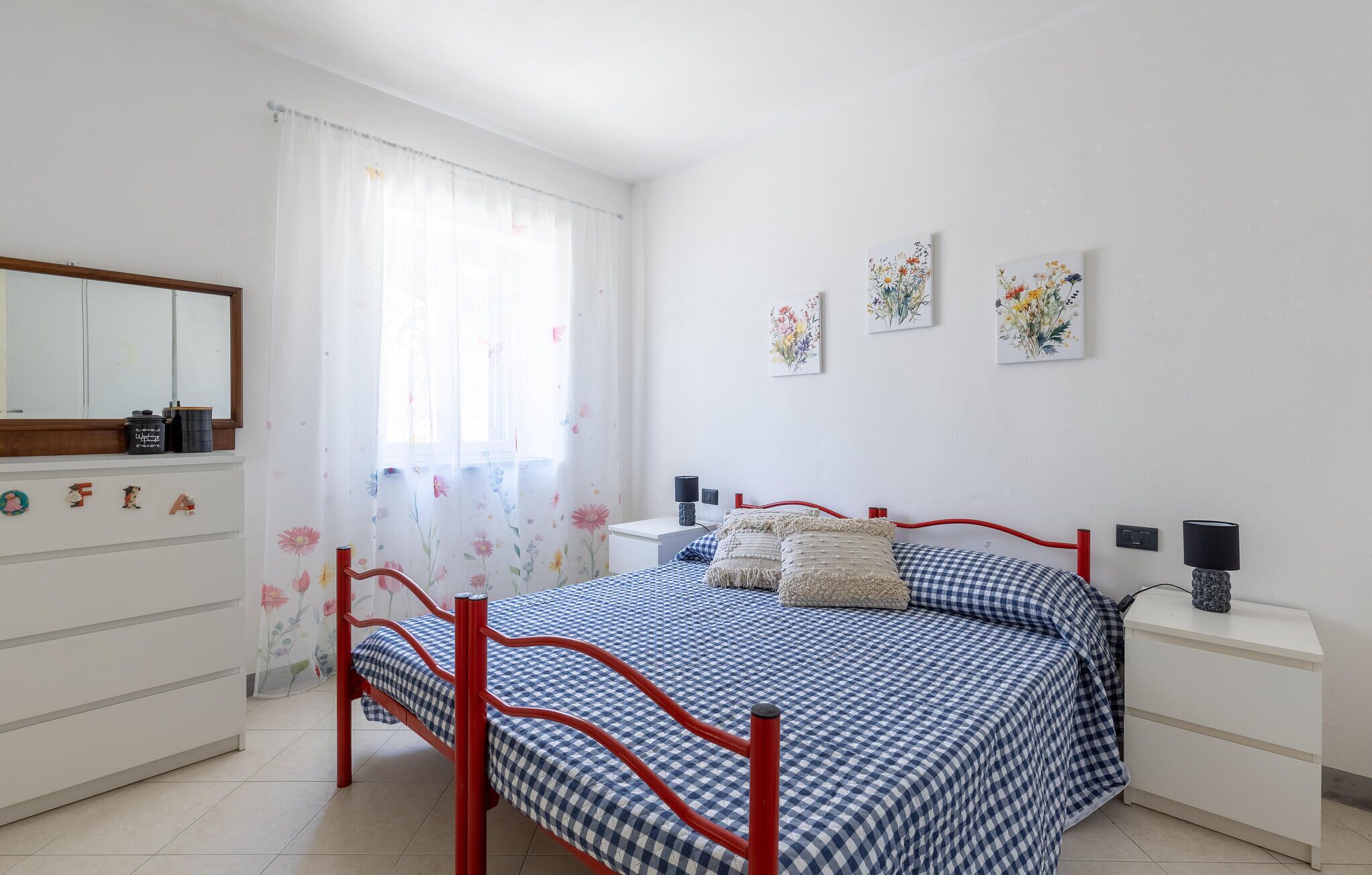 Une chambre à coucher avec un lit en métal rouge, une couvre-lit à carreaux, une commode blanche, une table de chevet et deux lampes fixées au mur.