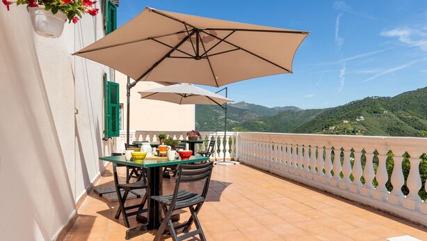 Una terraza con una mesa y sillas, una sombrilla color beige, y vistas a colinas verdes.