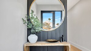 Ein modernes Badezimmer mit rundem Spiegel, einer Topfpflanze, einem Holzkonsolentisch und Blick auf ein Gebäude durch ein Fenster.