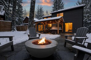 A snow-covered outdoor seating area with a fire pit, wooden chairs, and a building with a covered patio.