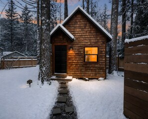 A wooden cabin with a snow-covered roof, a black door, and a lit window.