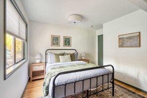 Un dormitorio con estructura de cama metálica, mesita de noche de madera, lámpara y dos cuadros en la pared.