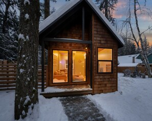 A wooden cabin with a warm interior, large windows, and a snow-covered landscape.