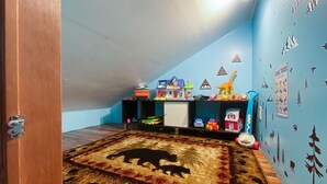A room with a sloped ceiling, a wooden floor, and a rug with animal silhouettes. There are shelves with toys and a wall with animal track decals.