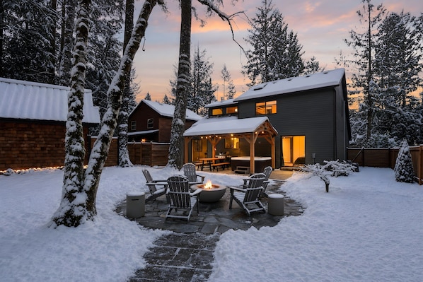 Une cour arrière enneigée avec un foyer extérieur, des sièges de jardin et une maison dotée d’un patio couvert.