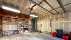 A garage with wooden shelving filled with tools and supplies, a concrete floor, and a ceiling with exposed beams and lighting fixtures.