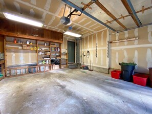 Eine Garage mit Holzböden, gefüllt mit Werkzeugen und Materialien, einem Betonboden sowie einer Decke mit freiliegenden Balken und Leuchten.