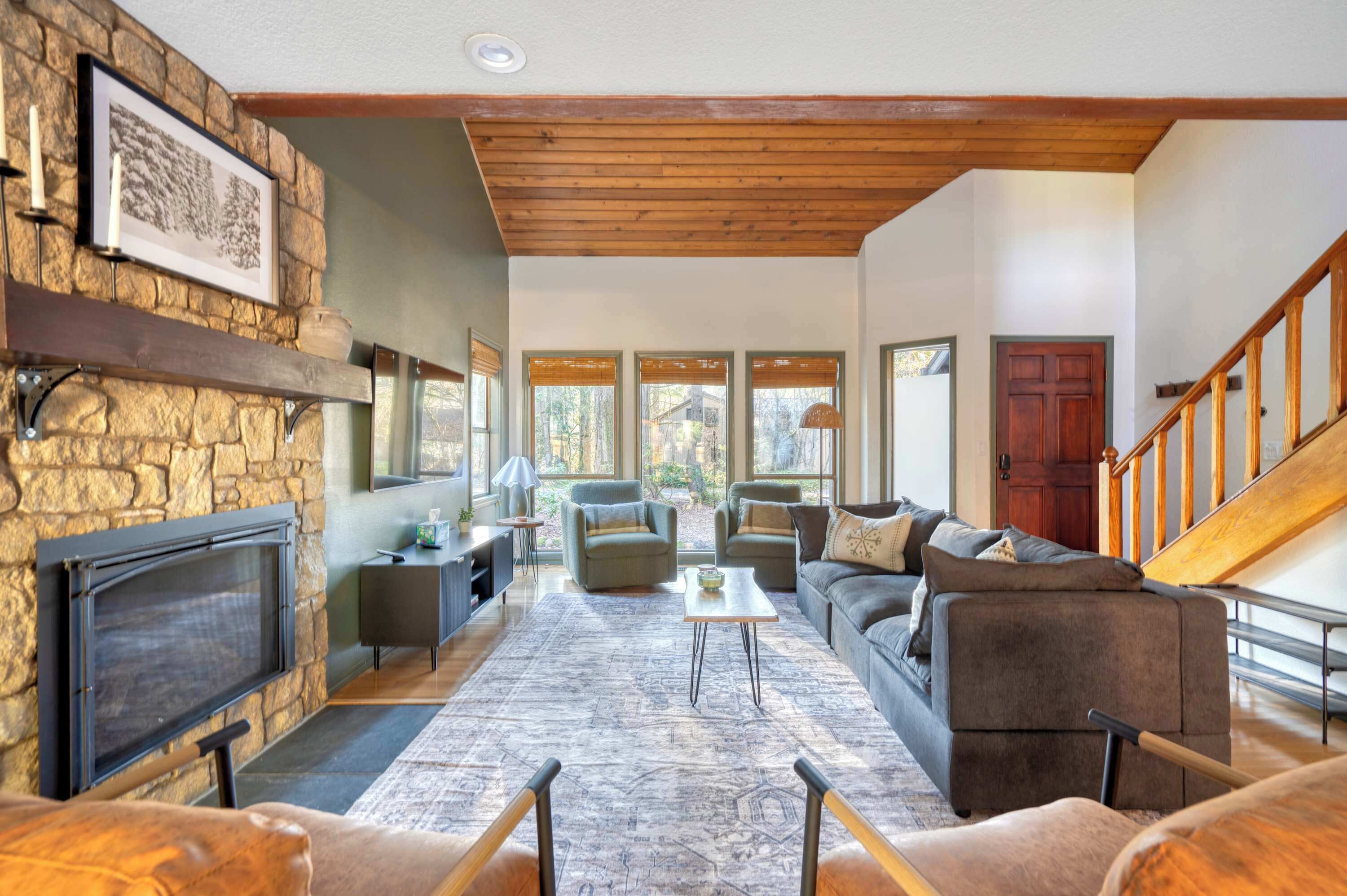 A spacious living room with a stone fireplace, large windows, and a wooden staircase.