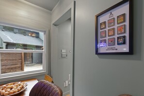A room with a large window, a framed collection of video game boxes on the wall, and a wooden table with a bowl of fruit.