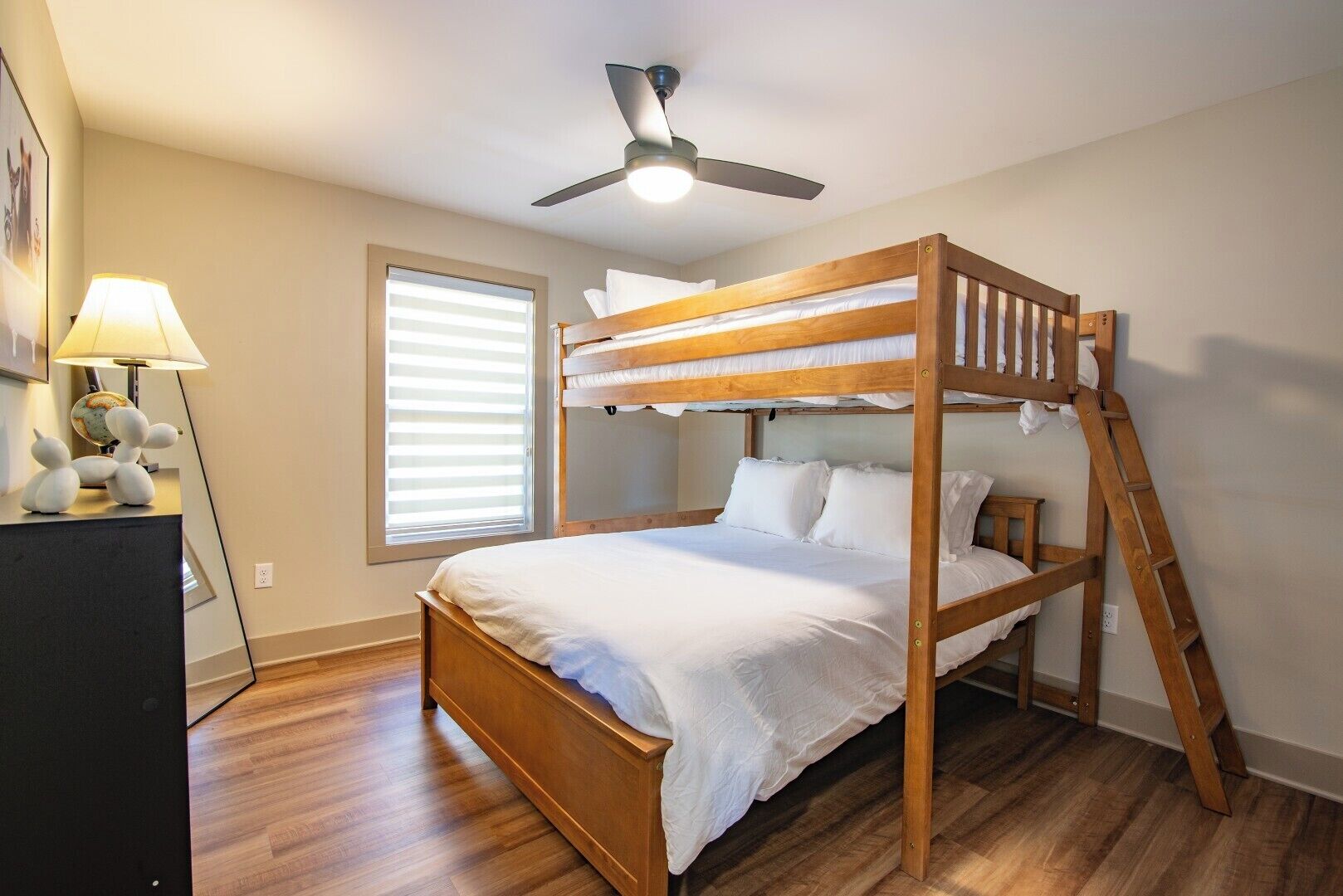 Habitación con literas, ventilador de techo, suelo de madera, escritorio con lámpara y ventana con persianas.
