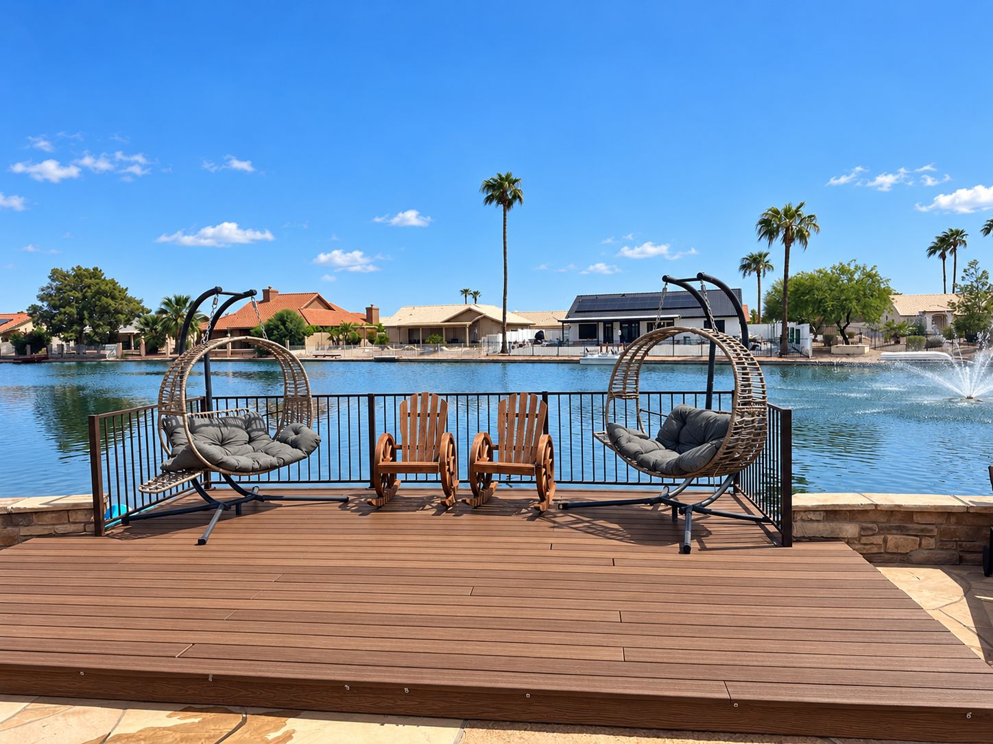 A wooden deck with two wicker chairs and a wooden table, overlooking a calm body of water with a fountain, surrounded by palm trees and buildings.