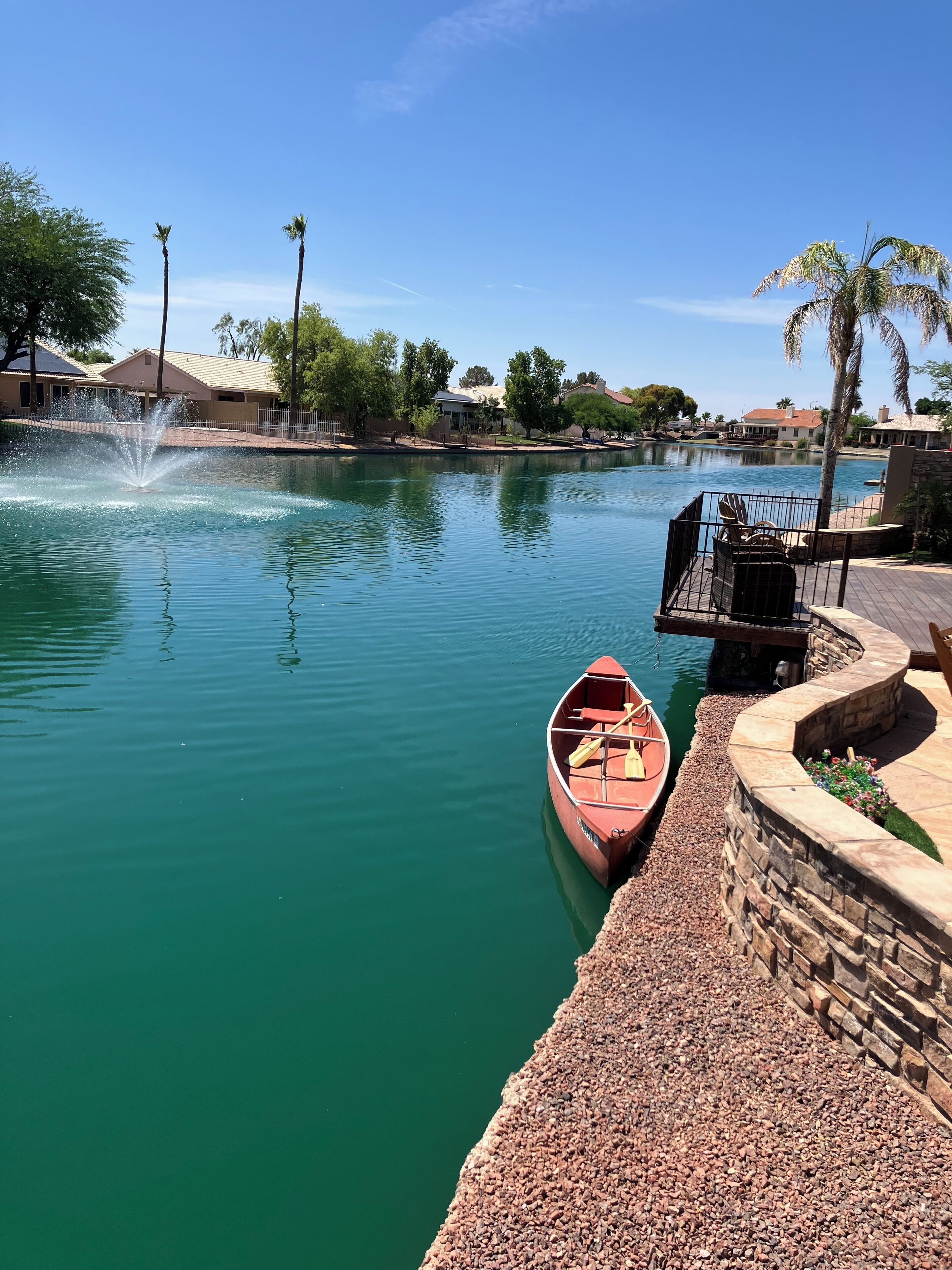 A calm water body with a small boat docked on the right, a fountain in the center, and a stone walkway along the edge.