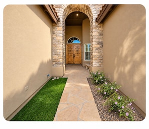 A pathway leading to a wooden door with a stone archway, a small garden with grass and flowers, and a window with shutters.