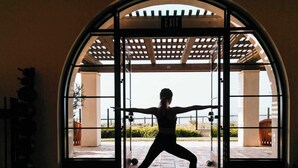 A person performing a yoga pose in a room with a large arched doorway, a ceiling with a grid pattern, and a view of an outdoor area with plants and a bench.