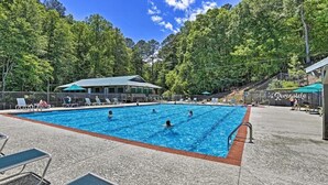 A swimming pool with multiple lanes, surrounded by lounge chairs and a covered seating area, set against a backdrop of dense trees.