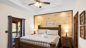 A bedroom with a wooden bed, two bedside tables with lamps, a ceiling fan, and a wooden wall with a 'kindness' sign.