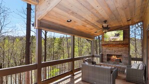 A covered outdoor deck with a stone fireplace, wicker furniture, and a view of the forest.