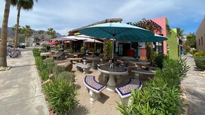 Un espace extérieur aménagé en terrasse, avec des tables et des bancs en pierre, entouré de verdure et de palmiers.