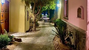 Un étroit sentier pavé, bordé de bâtiments aux murs roses et jaunes, qui mène à une rue bordée d’arbres.