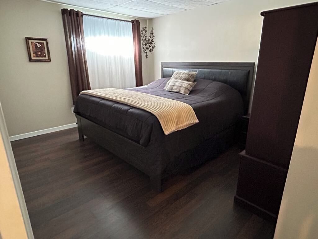 A bedroom with a dark-colored bed, a headboard, a window with curtains, and a framed picture on the wall.