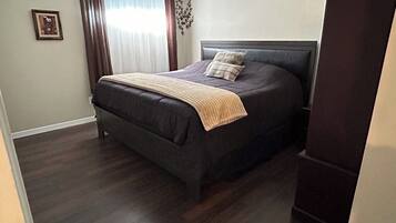A bedroom with a dark-colored bed, a headboard, a window with curtains, and a framed picture on the wall.