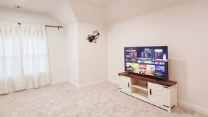 Une pièce spacieuse avec un grand téléviseur à écran plat posé sur un meuble télé en bois, des murs blancs et un sol recouvert de moquette.