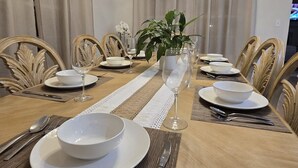 Une salle à manger avec une longue table en bois, dressée pour un repas, entourée de chaises à dossiers décorés. Un pot de fleurs est placé au centre de la table, et un lustre est suspendu au plafond.